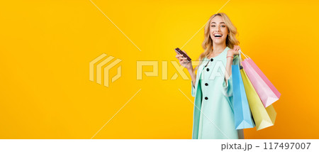 A young woman with blonde hair is smiling at the camera while holding colorful shopping bags in one hand and a phone in the other. She is wearing a light blue coat, panorama with copy space 117497007