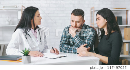 A doctor in a white coat is talking to a couple during a medical consultation in an office. The man is sitting with his head in his hands, while the woman is sitting next to him A doctor in a white coat is talking to a couple during a medical consultation in an office. The man is sitting with his head in his hands, while the woman is sitting next to him 117497025