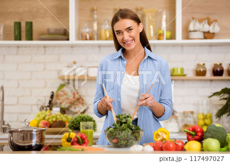A woman smiles while mixing a salad in her kitchen, surrounded by fresh vegetables and a pot on the counter. 117497027