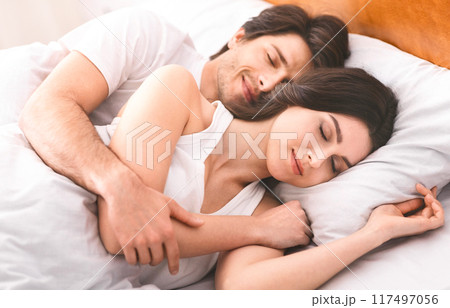 A couple sleeps peacefully in bed together at home. The woman is lying on her side, her head resting on a pillow, while the man is hugging her from behind. They both have their eyes closed 117497056
