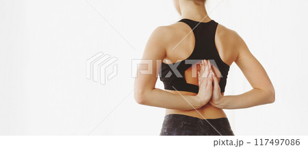 A woman is seen practicing yoga in her home, performing a challenging reverse prayer pose while wearing black sportswear during the morning, panorama with copy space 117497086