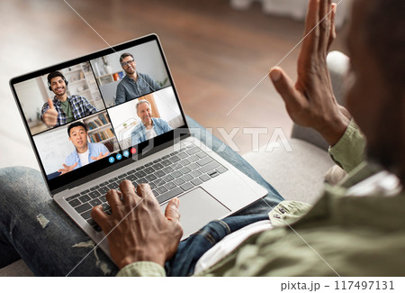 A man is sitting on a couch and participating in a video conference call with three other colleagues on his laptop. He is using his hand to gesture and is clearly engaged in the conversation 117497131