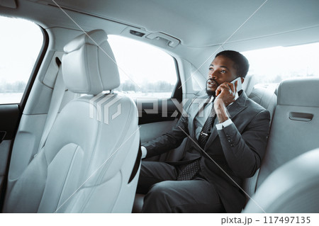 African American businessman in a gray suit is sitting in the back seat of a car and talking on a cell phone. He is looking out the window with a thoughtful expression on his face. African American businessman in a gray suit is sitting in the back seat of a car and talking on a cell phone. He is looking out the window with a thoughtful expression on his face. 117497135