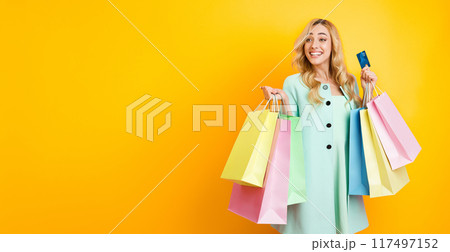 A happy woman with long blonde hair is standing in front of a yellow background. She is holding several shopping bags in one hand and a credit card in the other hand, copy space A happy woman with long blonde hair is standing in front of a yellow background. She is holding several shopping bags in one hand and a credit card in the other hand, copy space 117497152