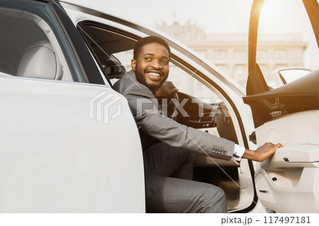 A smiling black businessman in a gray suit exits a white car on a sunny day. The car is parked on a street in an urban setting. He is likely heading to a meeting or appointment. A smiling black businessman in a gray suit exits a white car on a sunny day. The car is parked on a street in an urban setting. He is likely heading to a meeting or appointment. 117497181