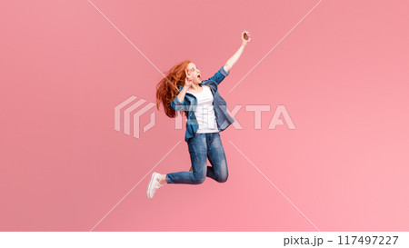 A spirited red-haired woman is caught mid-leap against a cheerful pink backdrop, her arm outstretched holding a smartphone as if capturing a moment of pure joy A spirited red-haired woman is caught mid-leap against a cheerful pink backdrop, her arm outstretched holding a smartphone as if capturing a moment of pure joy 117497227