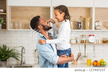 Little african girl and her cheerful father bonding at kitchen, daughter playing with dad nouse, empty space 117497237