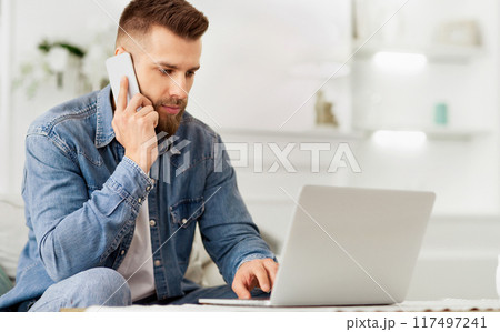 A man seated on the floor, multitasking by using a laptop and talking on a cell phone. 117497241