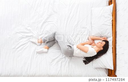 A woman is lying on a bed covered with crisp white sheets. She appears relaxed, with her eyes closed and hands resting by her side. The room is bright and minimalistic, top view A woman is lying on a bed covered with crisp white sheets. She appears relaxed, with her eyes closed and hands resting by her side. The room is bright and minimalistic, top view 117497284