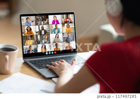 A woman in a red shirt participates in a large online video conference meeting on her laptop. Her hands are on the trackpad, and she is looking at the screen with a focused expression. 117497285