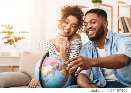 African American man and woman are sitting comfortably on a couch, attentively examining a globe placed between them. They seem engrossed in studying the geography displayed on the globe. 117497303