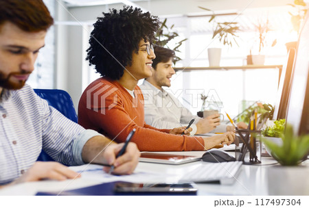 A diverse group of multiethnic men are seated around a desk, focused on a computer screen. They appear to be engaged in a collaborative work or discussion, with papers and notebooks scattered around. 117497304