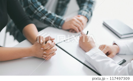 This image shows a close up of the hands of a patient and a doctor during a medical consultation. The patient has their hands clasped together on the table in front of them 117497309