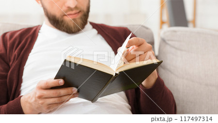 A man is seated on a couch, engrossed in reading a book. The man appears focused and relaxed, surrounded by a quiet atmosphere, cropped A man is seated on a couch, engrossed in reading a book. The man appears focused and relaxed, surrounded by a quiet atmosphere, cropped 117497314