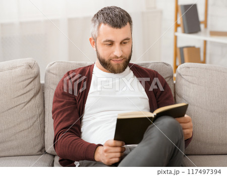 Bearded man is seated comfortably on a couch, engrossed in a book he is reading. The room is quiet as he flips through the pages, his focus solely on the words in front of him. 117497394