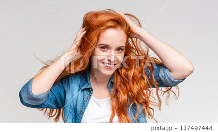 A cheerful, red-haired young woman is captured mid-movement as she runs her fingers through her hair, sporting a casual white t-shirt and denim jacket 117497448