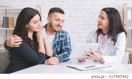 A young couple sits at a doctors office, listening intently as the doctor explains something on a tablet. The woman is leaning into the mans arm, and he is looking at the tablet with a smile 117497482