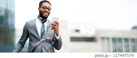 An African American businessman with his bicycle is using his phone. The mood is relaxed yet professional, capturing a modern lifestyle in an outdoor setting. 117497579