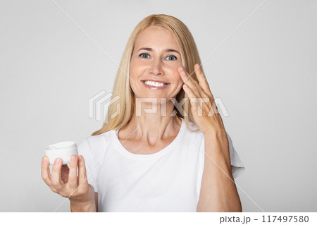 A woman with long blonde hair smiles as she applies cream to her face. She is wearing a plain white t-shirt and is standing in front of a simple grey backdrop. A woman with long blonde hair smiles as she applies cream to her face. She is wearing a plain white t-shirt and is standing in front of a simple grey backdrop. 117497580