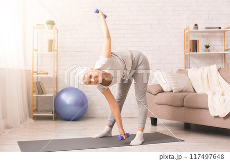 A smiling elderly woman is engaging in a fitness routine, holding a set of blue dumbbells while performing a side bend. She is standing on a yoga mat in a well-lit living room A smiling elderly woman is engaging in a fitness routine, holding a set of blue dumbbells while performing a side bend. She is standing on a yoga mat in a well-lit living room 117497648