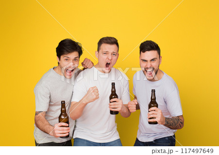 Three enthusiastic young men holding beer bottles cheer and shout together against a vivid yellow background, portraying friendship and celebration Three enthusiastic young men holding beer bottles cheer and shout together against a vivid yellow background, portraying friendship and celebration 117497649