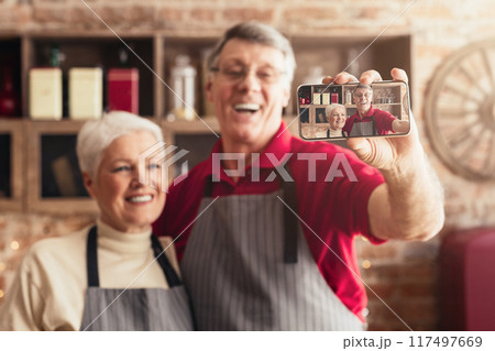 An older couple wearing aprons is taking a selfie in their kitchen. They are smiling and appear to be enjoying themselves while preparing a meal together. 117497669