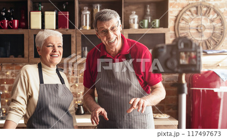 A senior couple films a cooking video in their home kitchen. The man is holding a red bell pepper and smiling at the camera, while the woman is standing behind him and smiling. 117497673