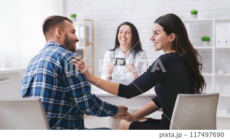 A couple sits facing each other in a doctors office, holding hands. The woman is smiling and placing her hand on the mans shoulder. 117497690