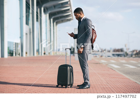 Black businessman in a suit checking his phone while standing with a rolling suitcase outside a modern building. 117497702