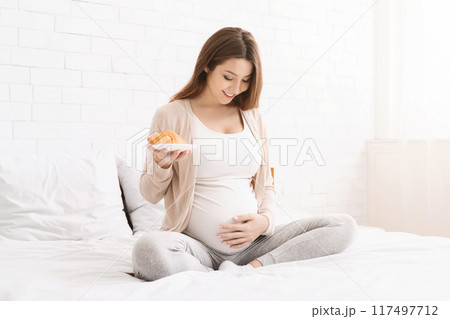 A pregnant woman is seated comfortably on a bed, holding a croissant in one hand while placing the other on her belly. The room is bright and filled with natural sunlight. 117497712