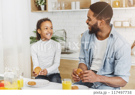 Sweet African American Girl And Her Dad Eating Cookies At Kitchen, drinking fresh orange juice, copy space 117497738