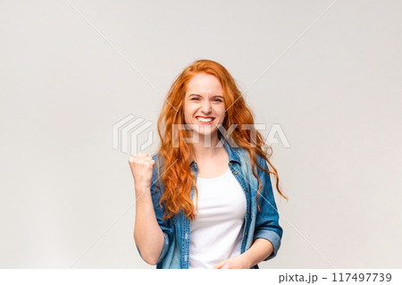 A radiant young woman with flowing red hair is captured mid-celebration, her fist pumped in victory and a beaming smile across her face, signaling triumph or good news A radiant young woman with flowing red hair is captured mid-celebration, her fist pumped in victory and a beaming smile across her face, signaling triumph or good news 117497739