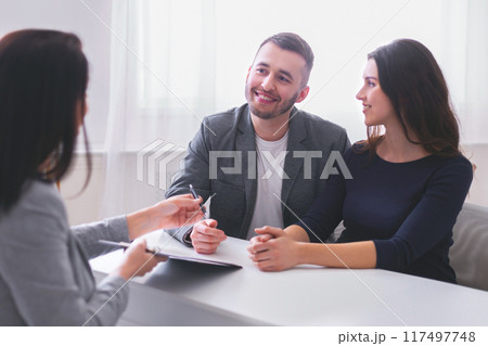 Happy couple sits across a desk from a financial advisor. The woman is smiling and looking at the advisor, while the man is looking at her. The advisor is pointing at a document on the desk 117497748