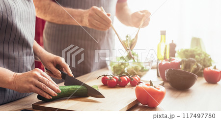 A couple is working together in the kitchen, preparing a fresh salad. The woman is using a knife to chop a cucumber on a wooden cutting board while the man is adding olive oil to a bowl of greens A couple is working together in the kitchen, preparing a fresh salad. The woman is using a knife to chop a cucumber on a wooden cutting board while the man is adding olive oil to a bowl of greens 117497780