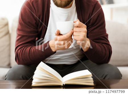 A man is seated on a couch, holding a cup in one hand and engrossed in reading a book. He appears relaxed and focused on the text, with a comfortable posture, cropped 117497799
