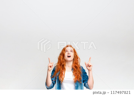 A young woman with vibrant red hair stands in front of a white backdrop, looking upwards with a look of surprise and excitement as she points at something unseen above her head, copy space A young woman with vibrant red hair stands in front of a white backdrop, looking upwards with a look of surprise and excitement as she points at something unseen above her head, copy space 117497821
