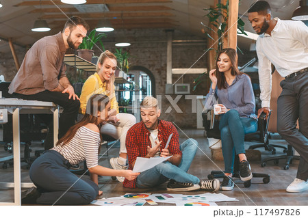 Diverse group of individuals sitting on the floor inside an office setting, engaged in a meeting or discussion. Diverse group of individuals sitting on the floor inside an office setting, engaged in a meeting or discussion. 117497826