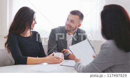 Couple sits at a table in an office, looking at documents with a financial advisor. Advisor holds up a sheet of paper while the couple looks attentive, suggesting they are in the midst of discussion 117497829