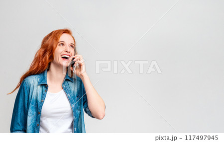 A woman with vibrant red hair is engaged in a phone conversation on her mobile device. She appears focused and animated as she communicates verbally through the phone, copy space 117497945