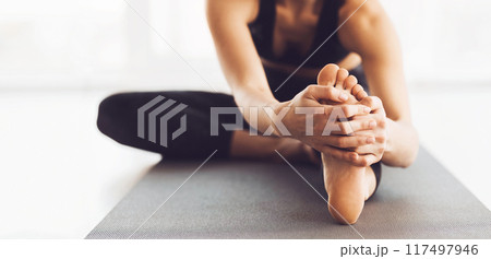 A woman is deeply engaged in her yoga practice, performing a seated forward bend stretch on a mat. The studio is filled with natural light, cropped A woman is deeply engaged in her yoga practice, performing a seated forward bend stretch on a mat. The studio is filled with natural light, cropped 117497946