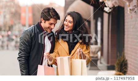 Woman showing man her purchases, guy looking into shopping bag outdoors Woman showing man her purchases, guy looking into shopping bag outdoors 117497958