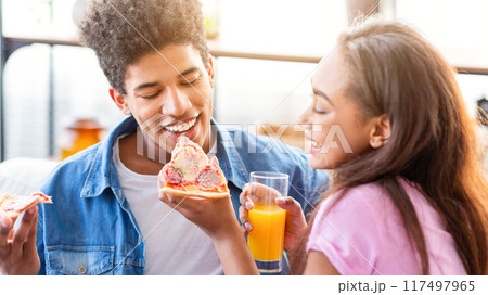 African American teen couple enjoys pizza together. The guy is biting into a slice of pizza and smiling. The girl is holding a glass of orange juice and watching him. They are both happy and relaxed. 117497965