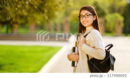 Asian young woman wearing glasses and a backpack is walking along a campus pathway, carrying books, and smiling on a sunny day. The park-like surroundings are lush and green 117498093