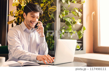 Small business development. Cheerful businessman working on laptop and talking on phone in cafe, free space 117498111