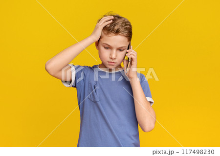 Fear and upset for mistake. Teenage boy talking on phone, holding hand on head in stress, orange studio background Fear and upset for mistake. Teenage boy talking on phone, holding hand on head in stress, orange studio background 117498230