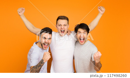Three young men are exuberantly celebrating, arms raised in triumph, against a vibrant orange background 117498260