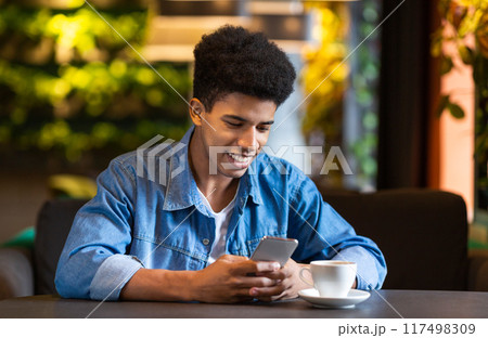 A young black guy is seated at a table, holding a cup of coffee in one hand and a cell phone in the other. He appears engrossed in his phone, possibly scrolling through messages or social media. A young black guy is seated at a table, holding a cup of coffee in one hand and a cell phone in the other. He appears engrossed in his phone, possibly scrolling through messages or social media. 117498309