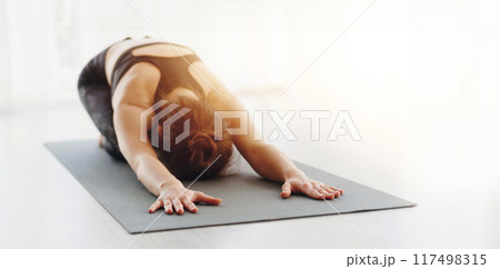 A woman is deeply engrossed in her yoga practice, performing the Childs Pose on a dark yoga mat. Surrounded by a softly lit studio environment, she stretches forward, copy space 117498315