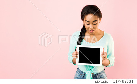A young black girl stands against a solid pink background during the day, holding a blank tablet in both hands while looking at it, mockup, copy space A young black girl stands against a solid pink background during the day, holding a blank tablet in both hands while looking at it, mockup, copy space 117498331