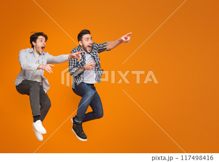 Two excited men pointing fingers away, jumping over orange background Two excited men pointing fingers away, jumping over orange background 117498416
